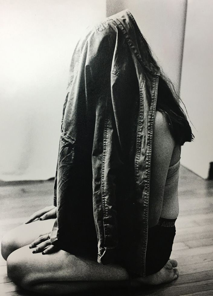 B&W photo: woman sitting on floor with head covered by a denim shirt/jacket