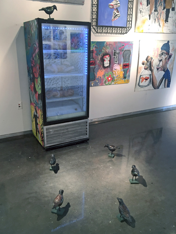 sculptural installation: a commercial display cooler, painted exterior, with babies on the shelves; a ring of birds sits on the ground in front of the cooler and one bird is perched on top