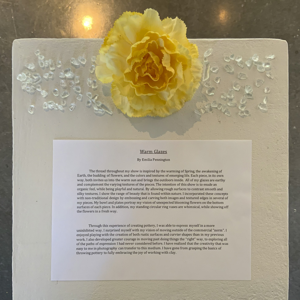 Pennington's artist statement on pedestal with yellow flower and glass shards