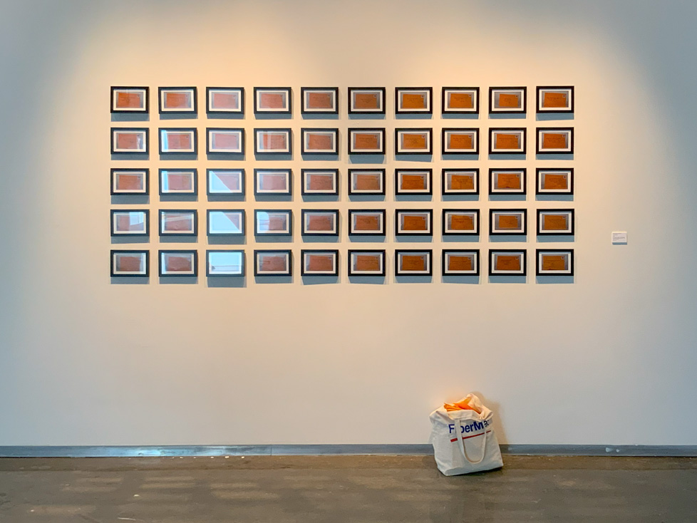 50 framed photos of envelopes mounted in a grid and a bag of envelopes on the floor below