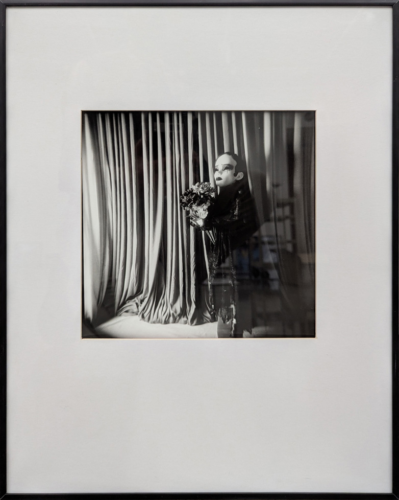 b/w photo of doll holding flowers, standing in front of curtains