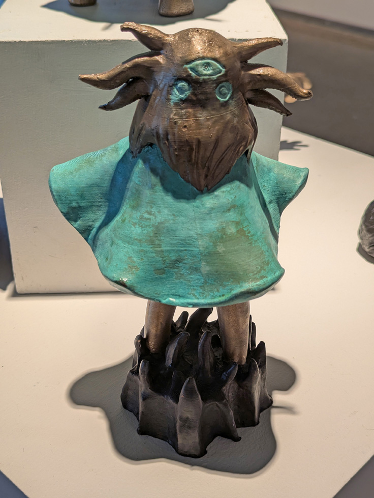 bronze cast of 3D-printed sculpture: bearded figure with three eyes and turqouise dress, standing in thick pointy base