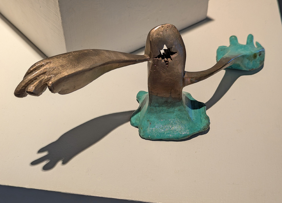 cast bronze sculpture of gumby-like figure with long arms, one weighted down with a large turquoise-colored hand