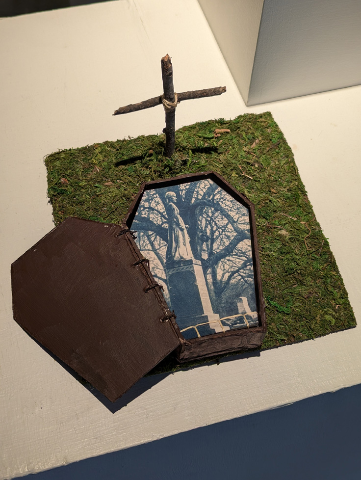 artist book in form of coffin on a grassy mat with stick cross containing cyanotype photos of graveyard