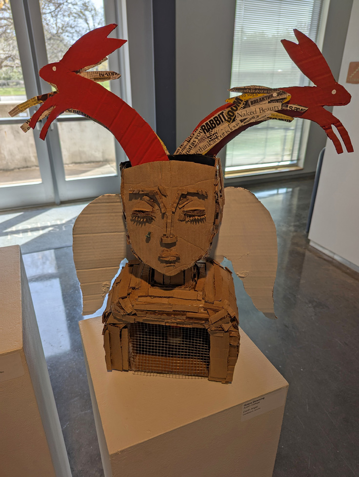 cardboard sculpture of crying angel with red rabbits springing from head
