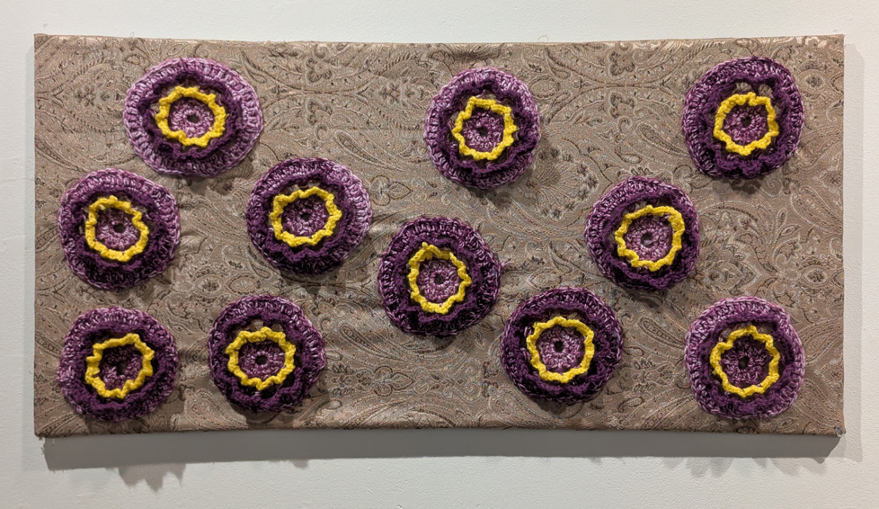 purple and yellow flowers made of yarn, attached to stretched taupe fabric
