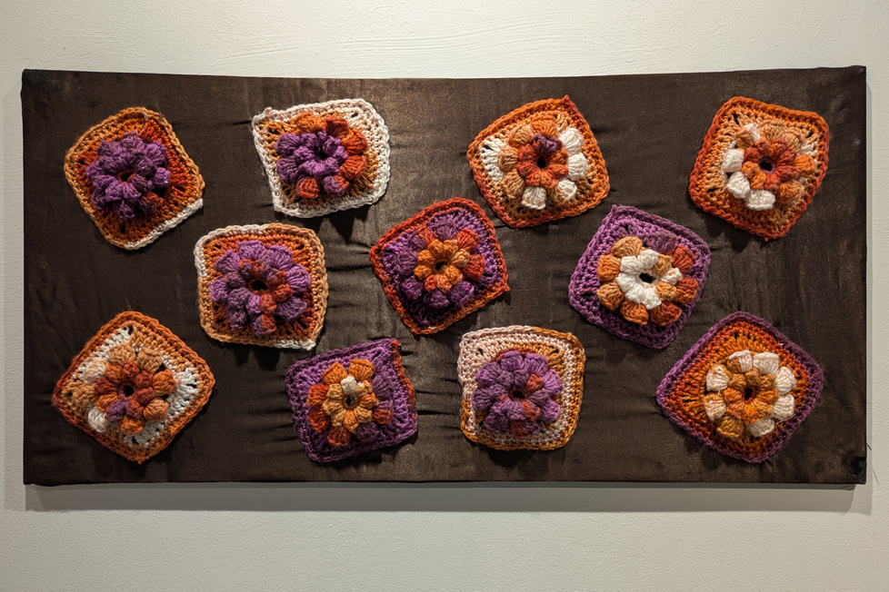 purple, orange, and white flowers made of yarn attached to stretched brown fabric with a sheen